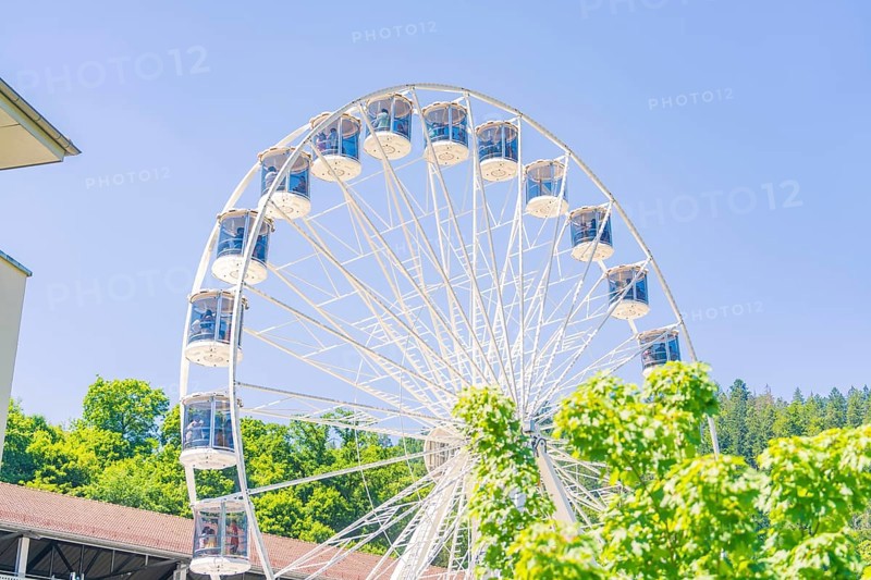 A white Ferris wheel turns in front of a building surrounded by green vegetation on a clear summer day, 950 years Calw, Germany