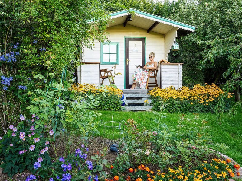 A woman in a dress sits relaxed on the small veranda of a garden shed and reads. Blooming summer flowers in the garden. Germany