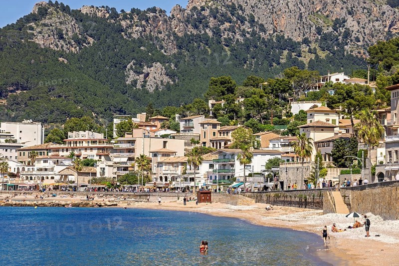 People sunbathing and bathing on the seashore at a idyllic coast village with houses at the boardwalk by the seabay and high mountains a sunny summer day, Port de Soller, Mallorca, Spain