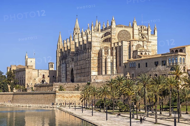 View at Palma Cathedral a famous Gothic Roman Catholic cathedral with the city walls and the Royal Palace of La Almudaina by a public park with palm trees and a pond a sunny summer day, Palma de Mallorca, Mallorca, Spain