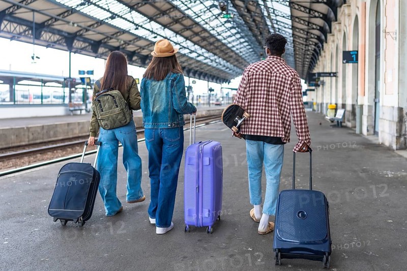 Three young tourists are walking away from camera in a train station with their suitcases and a skateboard, ready to start their vacation