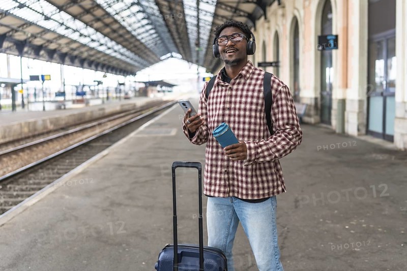 Young black male tourist waiting for his train while listening to music and holding a reusable water bottle, promoting sustainable travel