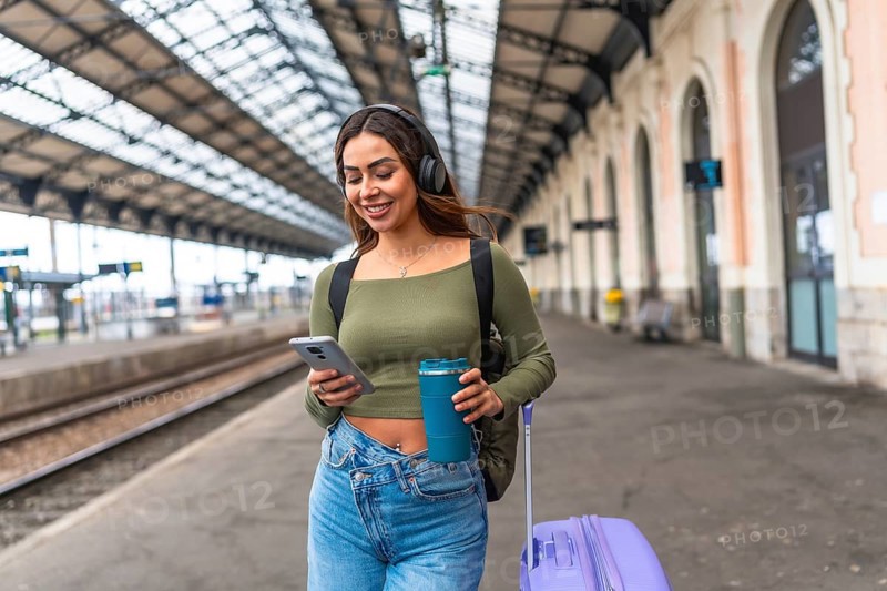 Young woman tourist walking in train station using smartphone and headphones while carrying reusable coffee cup and rolling suitcase