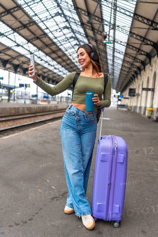 Tourist taking selfie with smartphone while waiting for train arrival at station platform, holding reusable coffee cup and carrying luggage