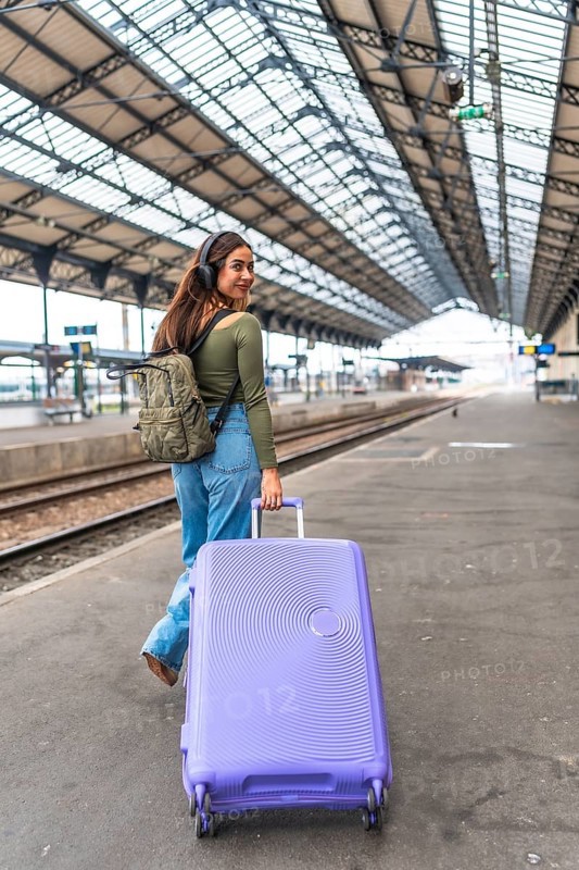 Tourist wearing headphones walking in train station pulling a purple suitcase and carrying a backpack