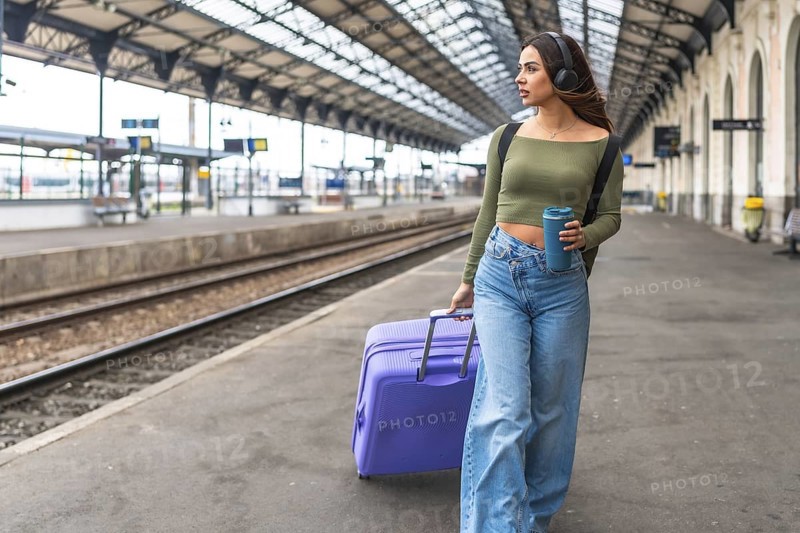 Tourist wearing headphones, holding reusable coffee cup and pulling suitcase while walking in train station