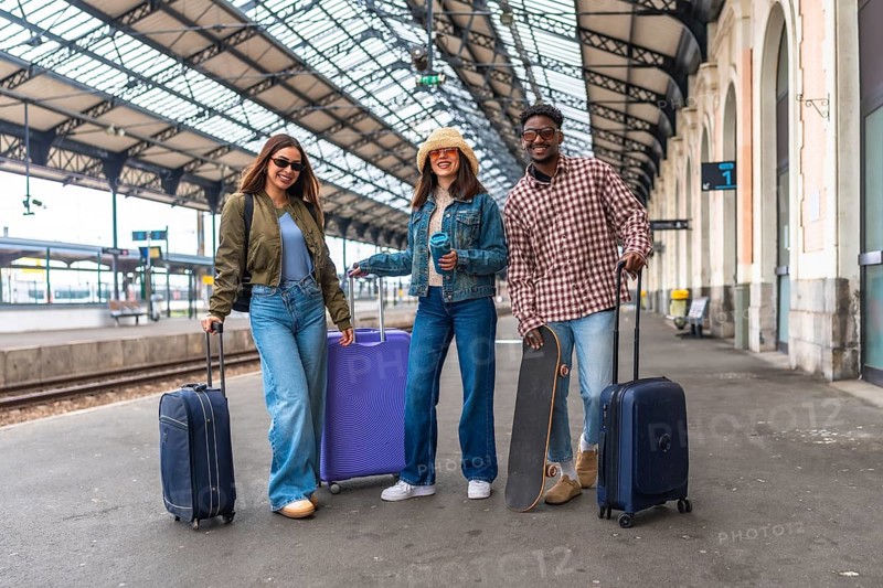 Happy tourists waiting at the railway station, surrounded by luggage and a skateboard, eagerly anticipating the train's arrival