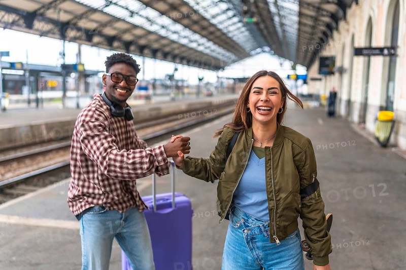 Young couple holding hands at train station, excited for their trip, with luggage and skateboard