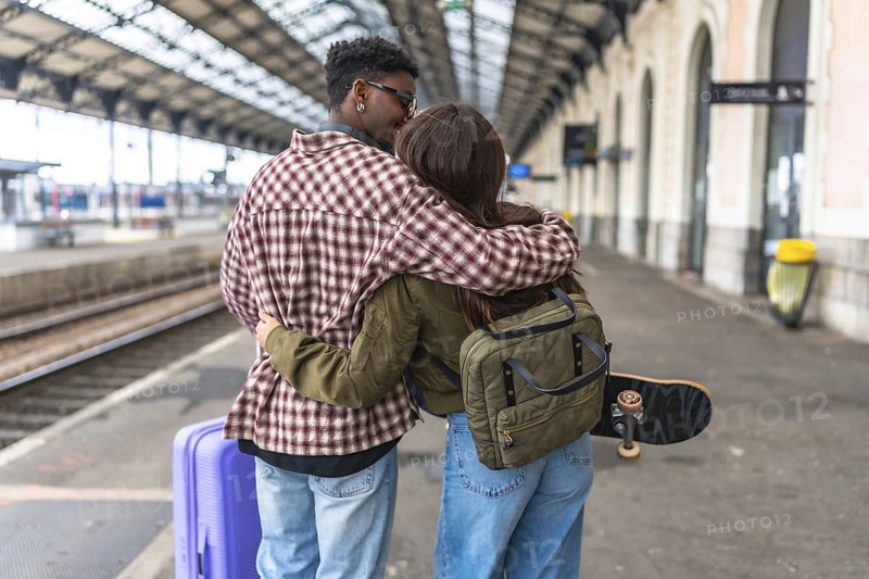 Young couple kissing and hugging on a train station platform, ready for their journey, with luggage and skateboard