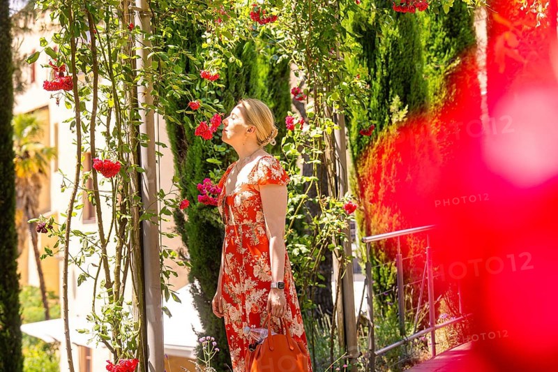 Woman in red dress smelling flowers on a plant path, surrounded by summer light, Botanical Garden, The Gardens of Trauttmansdorff Castle, Merano, South Tyrol, Italy