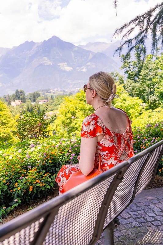 Woman in a red dress sitting on a bench and enjoying the view of the mountains, Botanical Garden, The Gardens of Trauttmansdorff Castle, Merano, South Tyrol, Italy