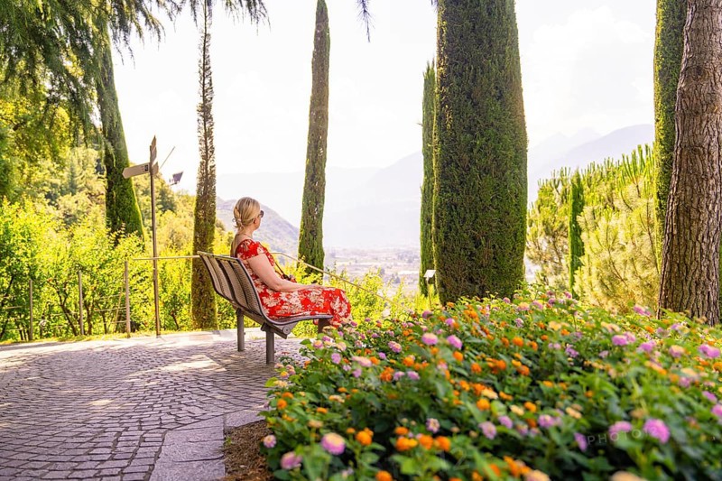 Woman in a red dress sitting on a bench overlooking a scenic view, Botanical Garden, The Gardens of Trauttmansdorff Castle, Merano, South Tyrol, Italy