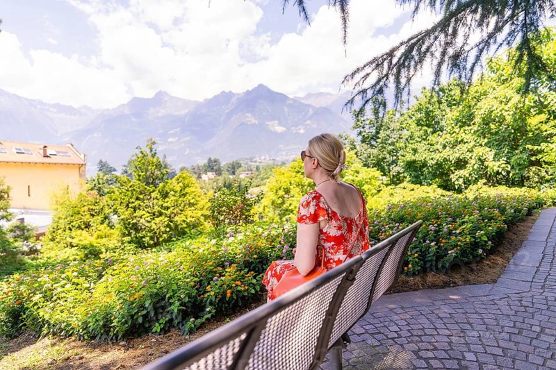 Woman in a red dress sitting on a bench with a view of mountains and nature, Botanical Garden, The Gardens of Trauttmansdorff Castle, Merano, South Tyrol, Italy