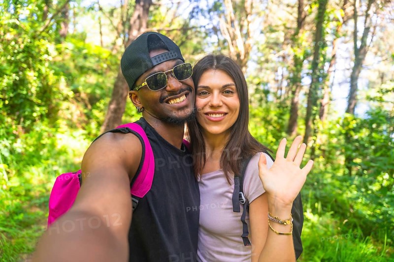 Interracial couple smiling and waving while taking a selfie during a vibrant hike in a lush, green forest filled with sunlight