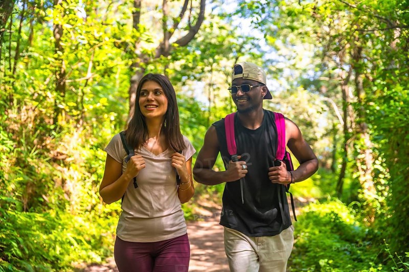 Interracial couple wearing backpacks walking in a forest trail, enjoying the beautiful nature surrounding them