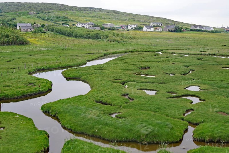 Green marshland with waterways and hills, behind a village under overcast skies, tidal landscape, cycle tour, Harris Hebrides, Scotland, Great Britain