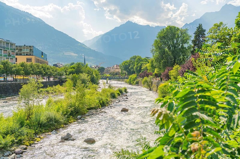 Panoramic view of a mountain river through a green landscape, in the background mountains under a blue sky, Merano, Italy