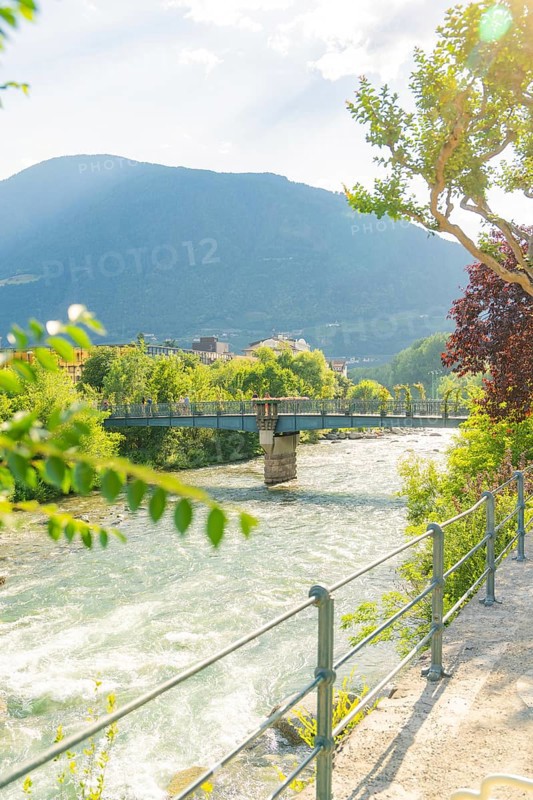 River with bridge, surrounded by green nature and mountains in sunlight, Merano, Italy