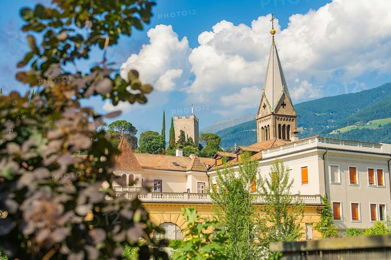 Historic building with church tower and picturesque mountains in the background under a cloudy sky, Merano, Italy