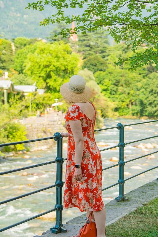A woman in a flowered dress looks at a flowing river on a sunny day, Merano, Italy