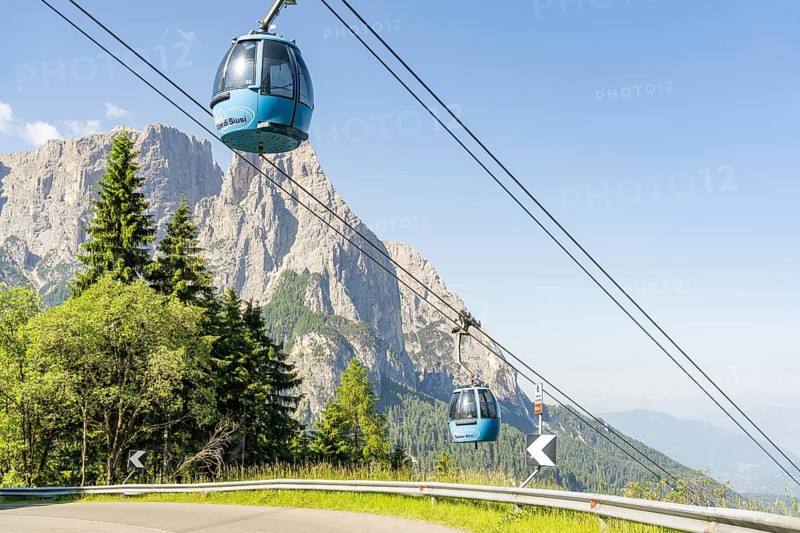 Two blue cable car gondolas hovering over trees in front of a rocky mountain range, Alpe di Siusi, Dolomites, South Tyrol, Italy
