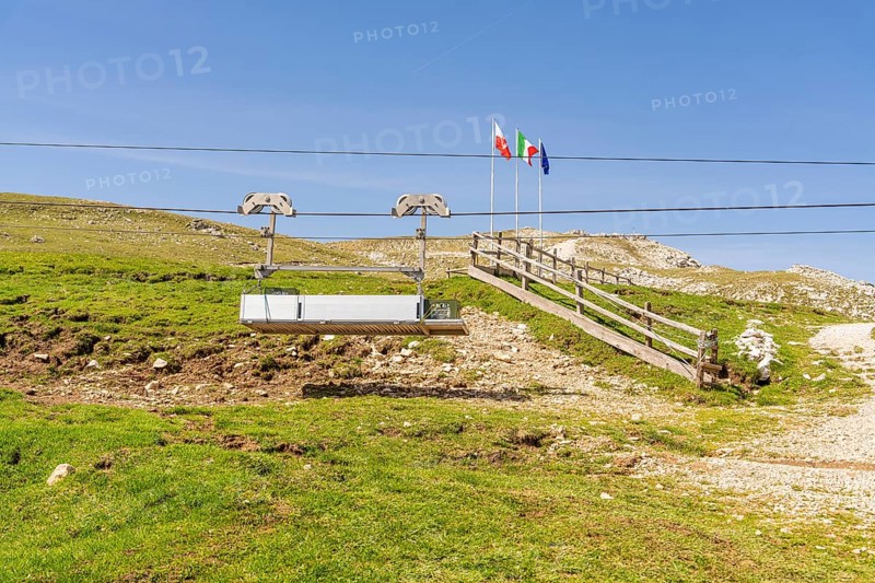 Chairlift in front of mountain landscape with flags and green slope under clear blue sky, Alpe di Siusi, Dolomites, South Tyrol, Italy