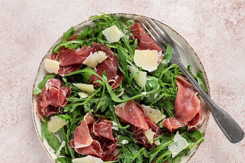 Fresh arugula salad with prosciutto and grated parmesan cheese, on a plate, homemade, no people