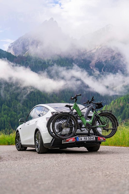 A car with bicycles drives along a road against a dramatic mountain backdrop and clouds, electric car on holiday, Dolomites, South Tyrol, Dolomites, Italy