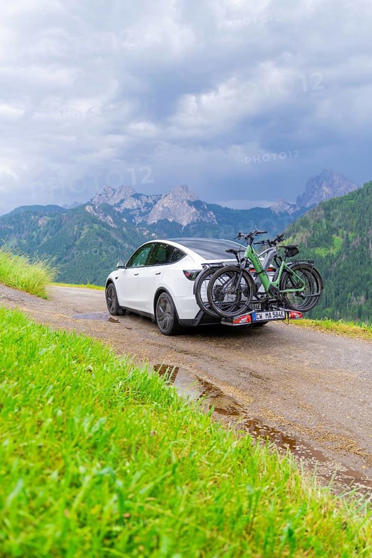 White car with bicycles on mountainous road, surrounded by green landscape and cloudy sky, electric car on holiday, Dolomites, South Tyrol, Dolomites, Italy