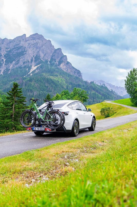 White car with bicycles on a road in a mountainous, green landscape with cloudy sky, electric car on holiday, Dolomites, South Tyrol, Dolomites, Italy
