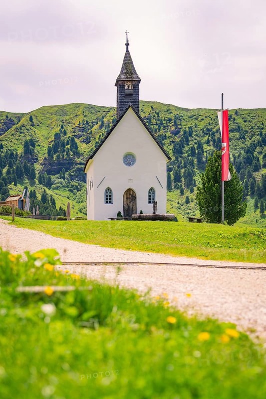 White church stands idyllically on a green meadow with mountain backdrop and flag, Alpe di Siusi, South Tyrol, Dolomites, Italy