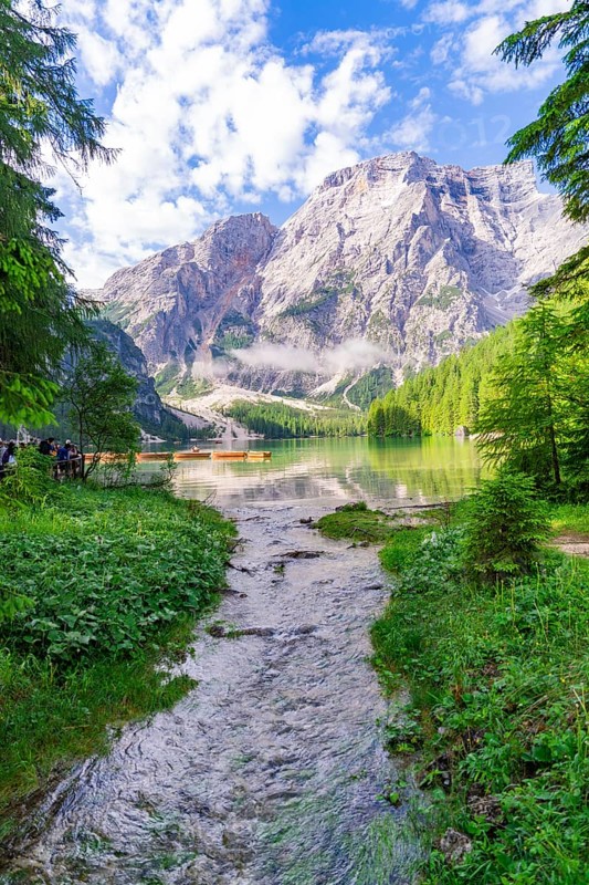 A clear mountain lake surrounded by impressive mountains and dense forest under a partly cloudy sky, Pragser Wildsee, South Tyrol, Dolomites, Italy