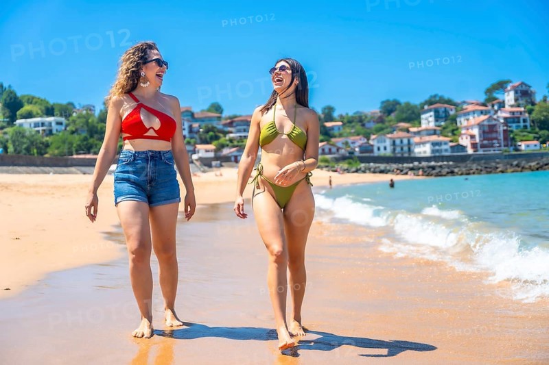 Two cheerful young women wearing bikinis and sunglasses are enjoying a walk on a sunny beach, creating a perfect summer vacation scene