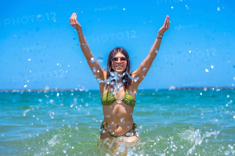 Young woman wearing sunglasses and a green bikini is raising her arms and splashing water in the sea, enjoying a sunny summer day at the beach