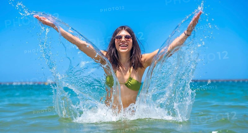 Young woman wearing stylish sunglasses and a vibrant bikini joyfully splashing in the clear ocean on a sunny summer day