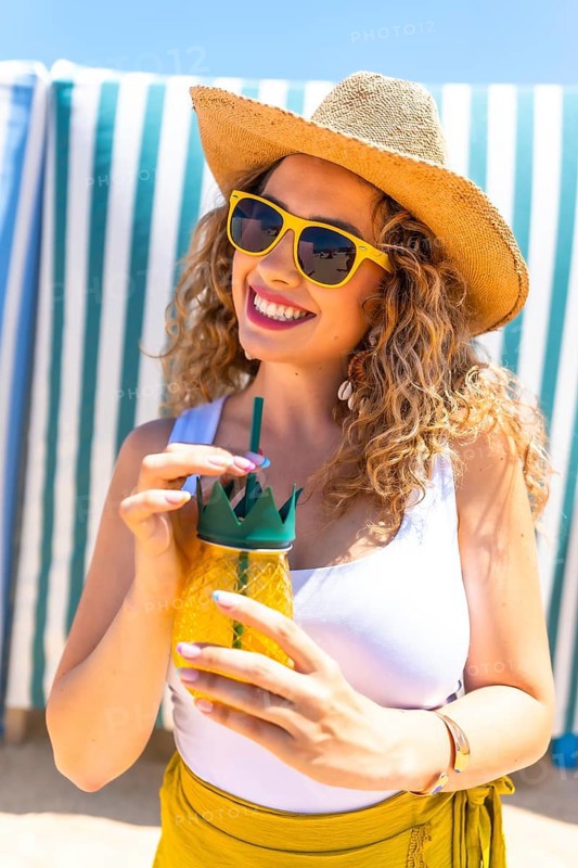 Young curly blonde woman wearing sunglasses and straw hat drinking juice at beach bar, enjoying summer vacation