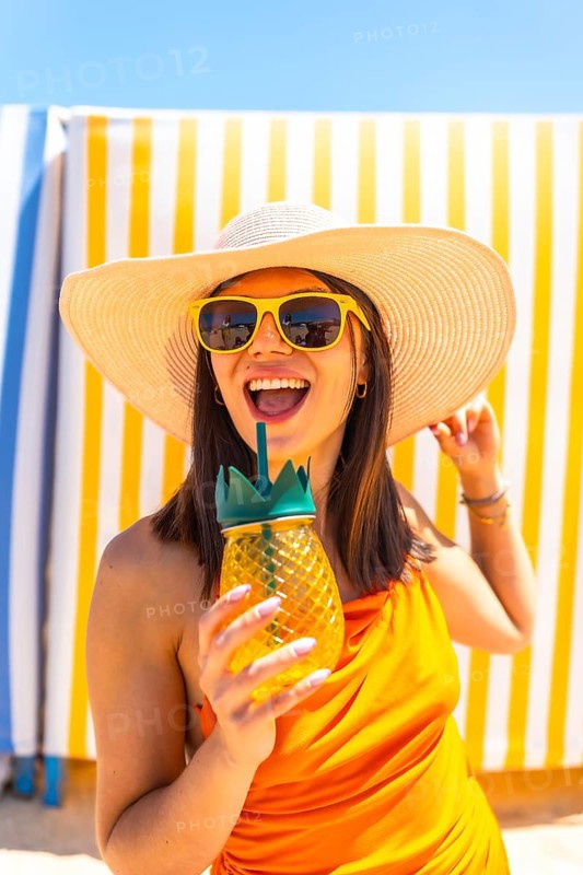 Young woman enjoying a refreshing pineapple cocktail while relaxing on the beach, wearing stylish sunglasses and a sun hat under the vibrant sky