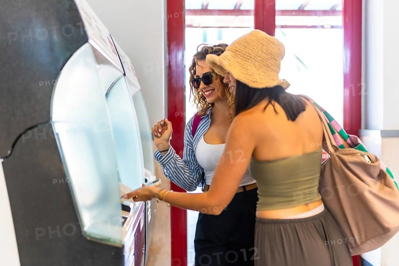 Two happy female tourists buying train tickets at automatic ticket machine inside station building