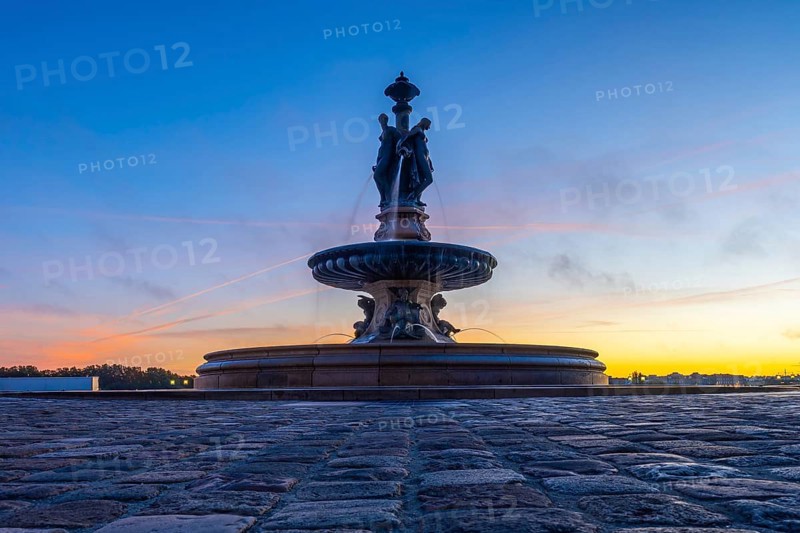 The three graces fountain in bordeaux, france, stands majestically against a vibrant sunrise sky, its waters flowing gracefully as dawn breaks over the city