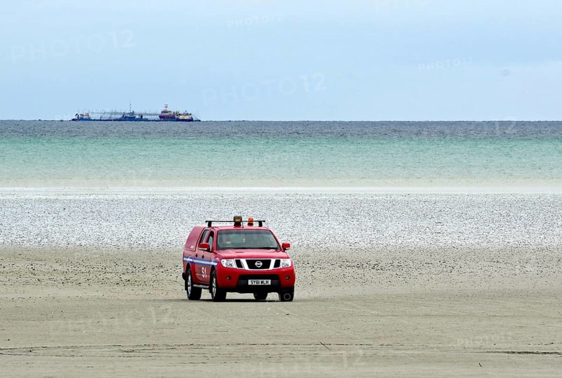 Red off-road vehicle on a sandy beach with sea and ships in the background under a cloudy sky, air traffic control, runway, tide, Isle of Barra, Hebrides, Scotland, Great Britain