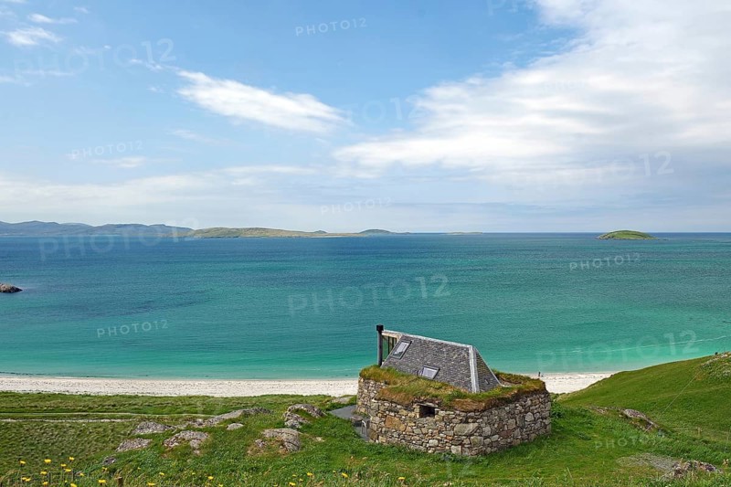 Stone cottage with grassy roof overlooking the vast sea and green hills under a blue sky, Eriksay, Hebrides, Scotland, United Kingdom
