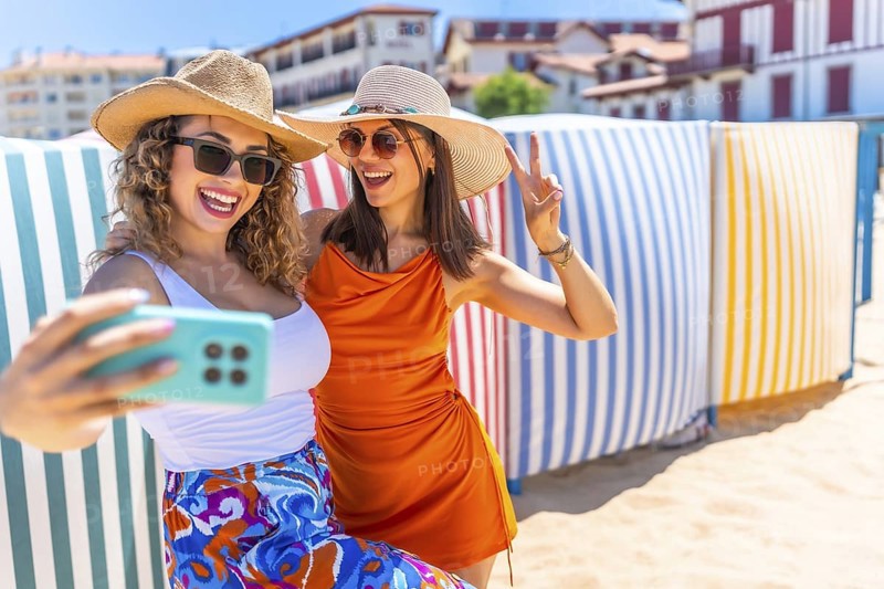 Two cheerful women wearing summer hats and sunglasses are taking a selfie with a smartphone on a sunny beach, gesturing peace signs in front of vibrant beach huts