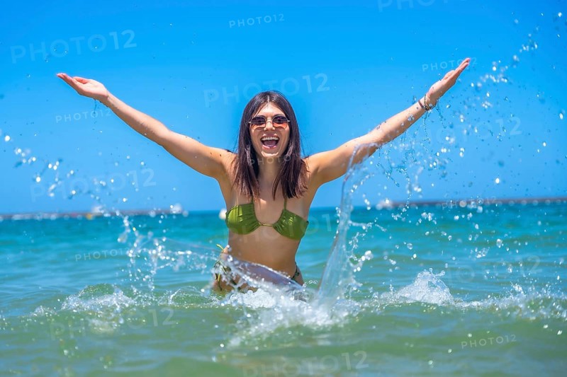 Young woman wearing sunglasses and a bikini is raising her arms and splashing water in the sea, enjoying her vacation