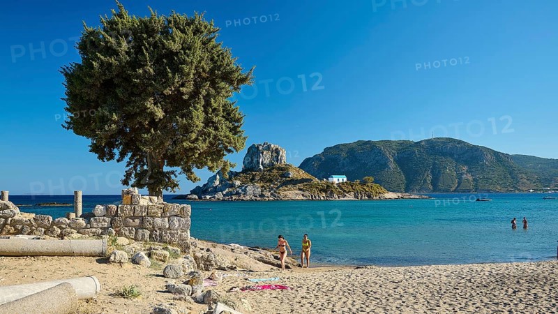 Family on the beach overlooking a green island and clear blue waters, Agios Stefanos Beach, Agios Stefanos Basilica, Agios Nikolaos Chapel, Kastri Island, Kos, Dodecanese, Greek Islands, Greece
