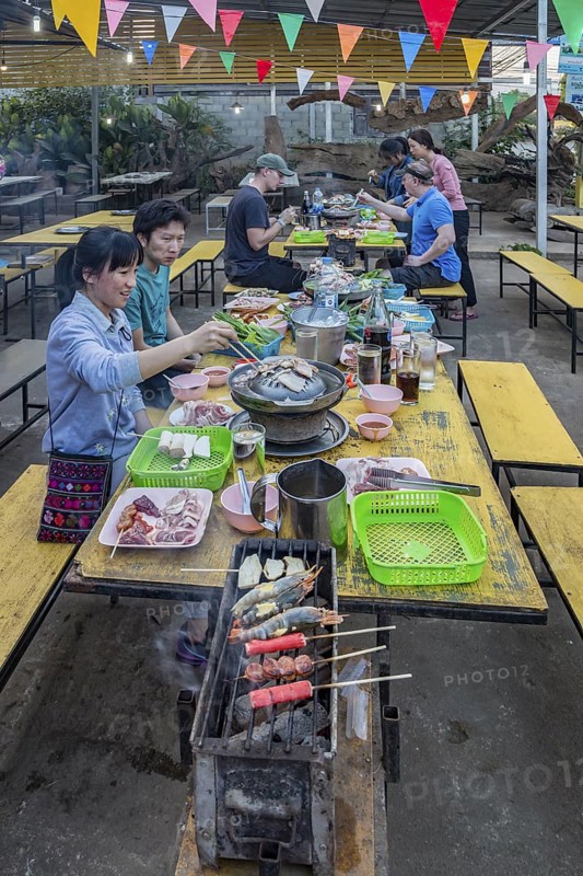 Thai man and woman cooking on a charcoal hot pot and grill at their table in a buffet style outdoor restaurant in Chiang Rai province of Northern Thailand