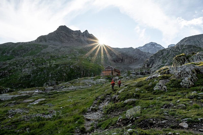 Mountaineer in front of mountain hut Elberfelderhütte with rocky mountain landscape, sun star in the morning, in the upper Gössnitz valley, Wiener Höhenweg, Schober group, Hohe Tauern National Park, Carinthia, Austria