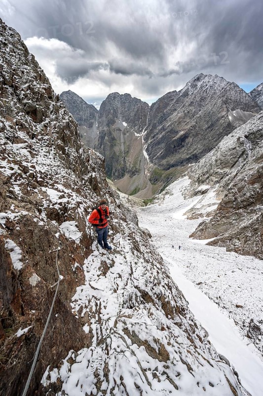 Mountaineer on a rope-secured path in a steep rocky mountain landscape with fresh snow in summer, behind mountain peaks Großer Friedrichskopf and Georgskopf, ascent to Hornscharte, Schobergruppe, Hohe Tauern National Park, Carinthia, Austria