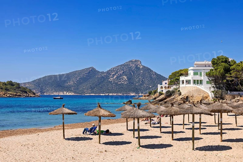 People sunbathing on a sandy beach with parasols by the Mediterranean Sea with white houses on a rocky coast, Sant Elm, Mallorca, Spain