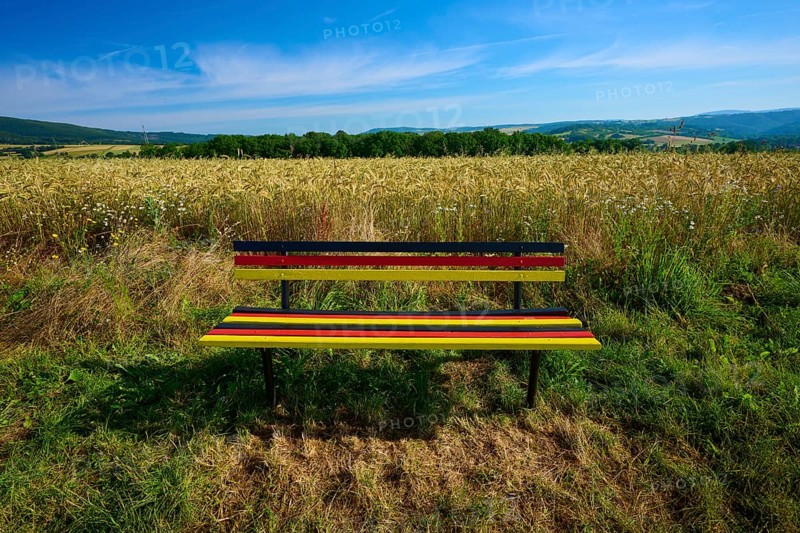 Bench in German colours with a wide view of a golden grain field and blue sky, summer, Germerode, Geo nature park Park Frau-Holle-Land, Hoher Meissner, Hesse, Germany