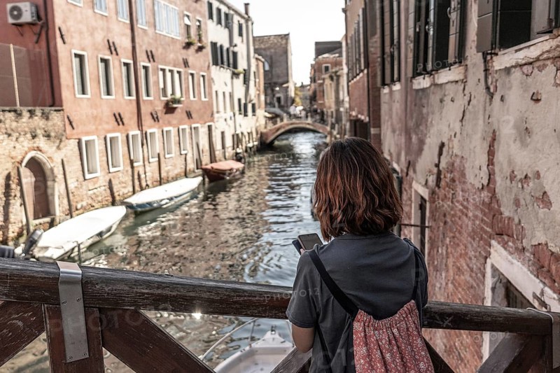 Young woman tourist with backpack using smartphone and enjoying view of picturesque canal, bridge, and historic buildings in venice, italy